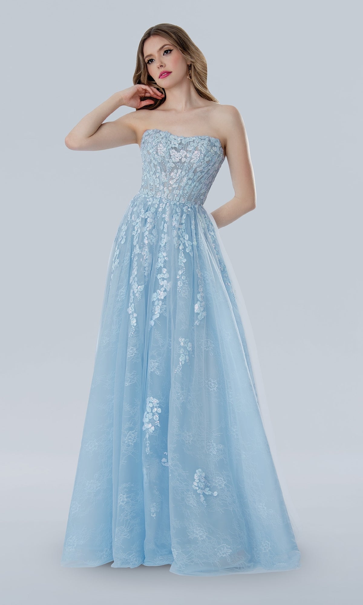 A woman stands against a plain light gray background wearing the Stella Couture 25108, a long strapless light blue prom dress with delicate floral lace embroidery.